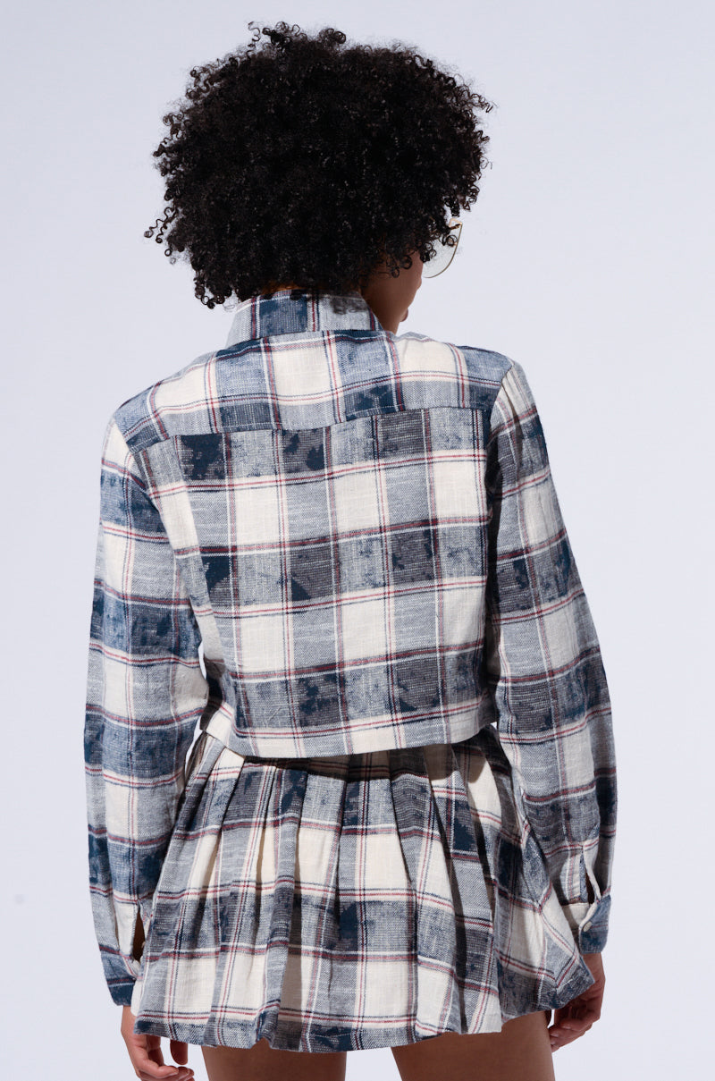 A person with curly hair is shown from the back wearing a BELLA PLAID BUTTON DOWN SHIRT in blue and white, made from 100% cotton, along with a matching Bella Plaid Pleated Mini Skirt. The slightly faded shirt is tucked into the pleated skirt, all set against a plain, light-colored background. 2