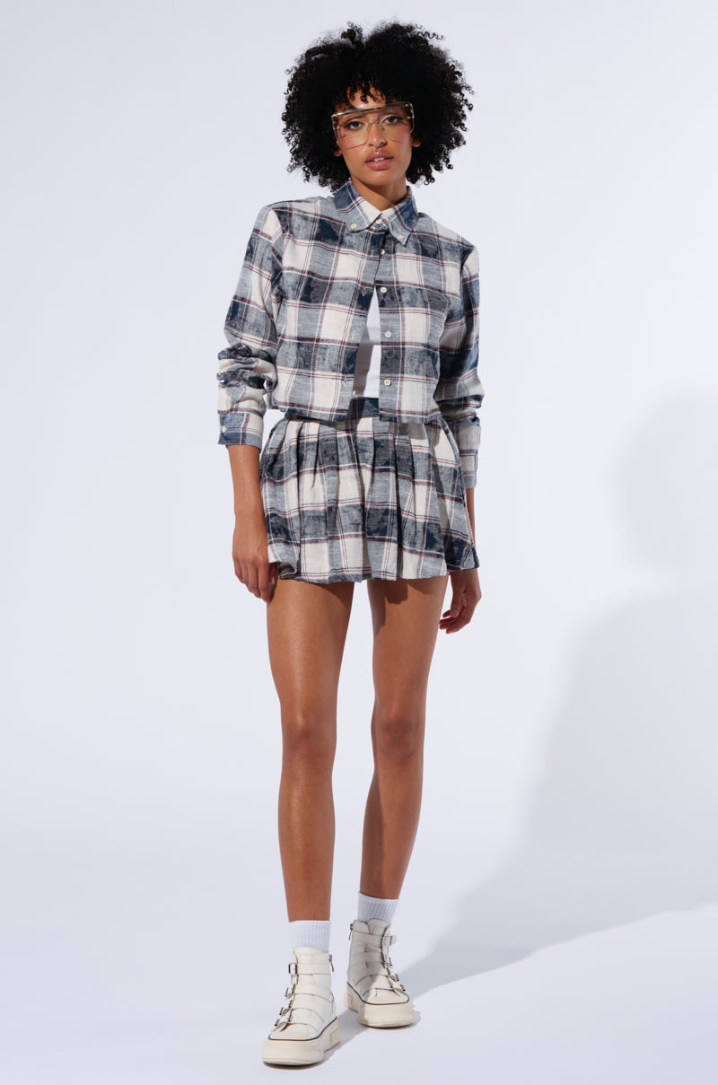 A person with short curly hair is wearing a checked, long-sleeve Bella Plaid Button Down Shirt and a Bella Plaid Pleated Mini Skirt. They are also wearing glasses, white socks, and white high-top sneakers. The background is plain and well-lit. 4