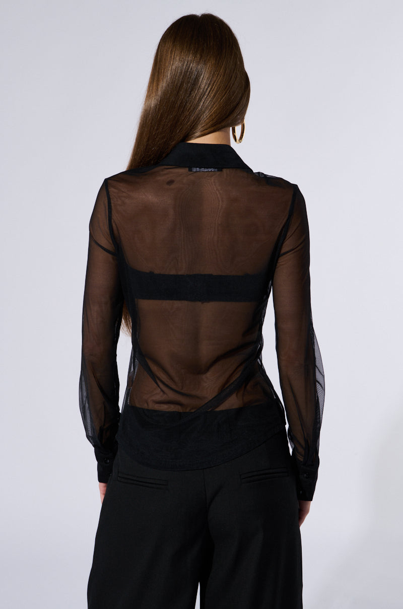 A woman is seen from behind, wearing a SEALED LIPS BUTTON DOWN BLOUSE with long sleeves and sheer mesh fabric paired with dark pants. The transparent blouse subtly reveals a black undergarment underneath. A rhinestone lip brooch adds a sparkle to her ensemble. Her long, straight hair cascades down her back against the plain white background.