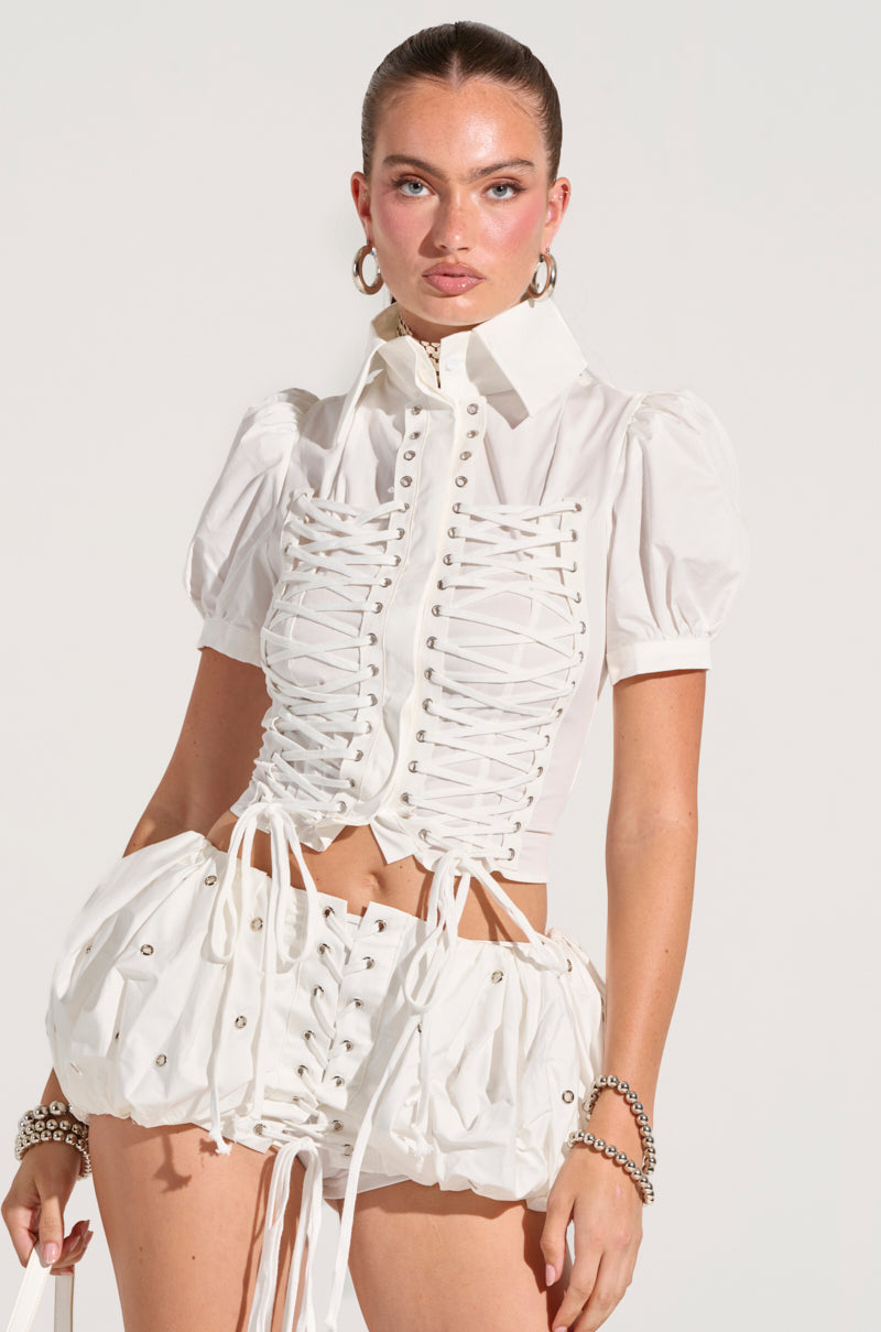 A person is styled in the PRETTY PLEASE BUTTON DOWN BLOUSE, a white, lace-up corset-style top made from a semi-stretchy cotton blend. It pairs effortlessly with matching bubble shorts and is adorned with puffed sleeves. The look is completed with hoop earrings and bracelets, all set against a plain, light background.