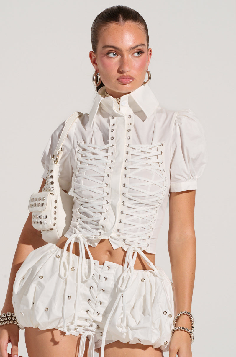 A woman stands wearing the chic PRETTY PLEASE BUTTON DOWN BLOUSE featuring lace-up paneling crafted from a semi-stretchy cotton blend. She carries a small studded white purse, her hair is pulled back, and she accessorizes with hoop earrings and a bracelet. The backdrop is plain.