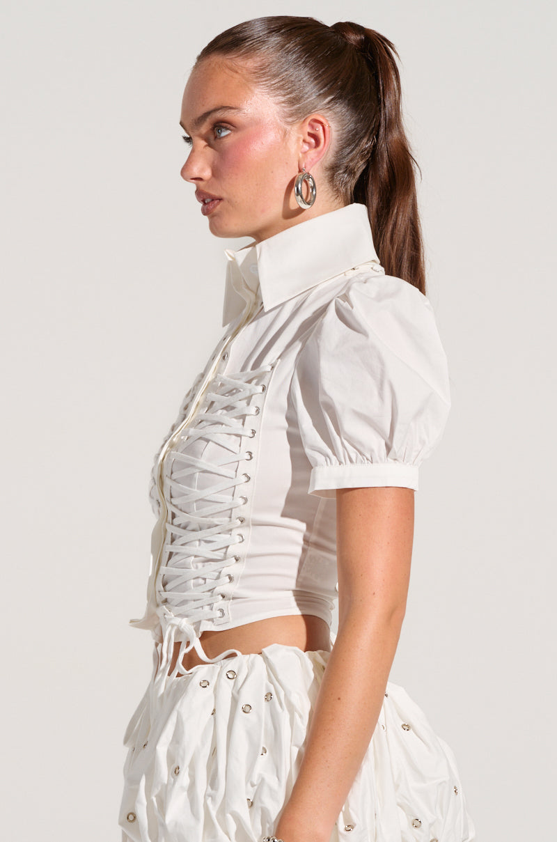 A person with long brown hair tied in a ponytail is wearing the PRETTY PLEASE BUTTON DOWN BLOUSE, which has lace-up paneling at the front, paired with a skirt made of semi-stretchy cotton blend featuring a subtly textured pattern. They are looking to the left against a plain background.
