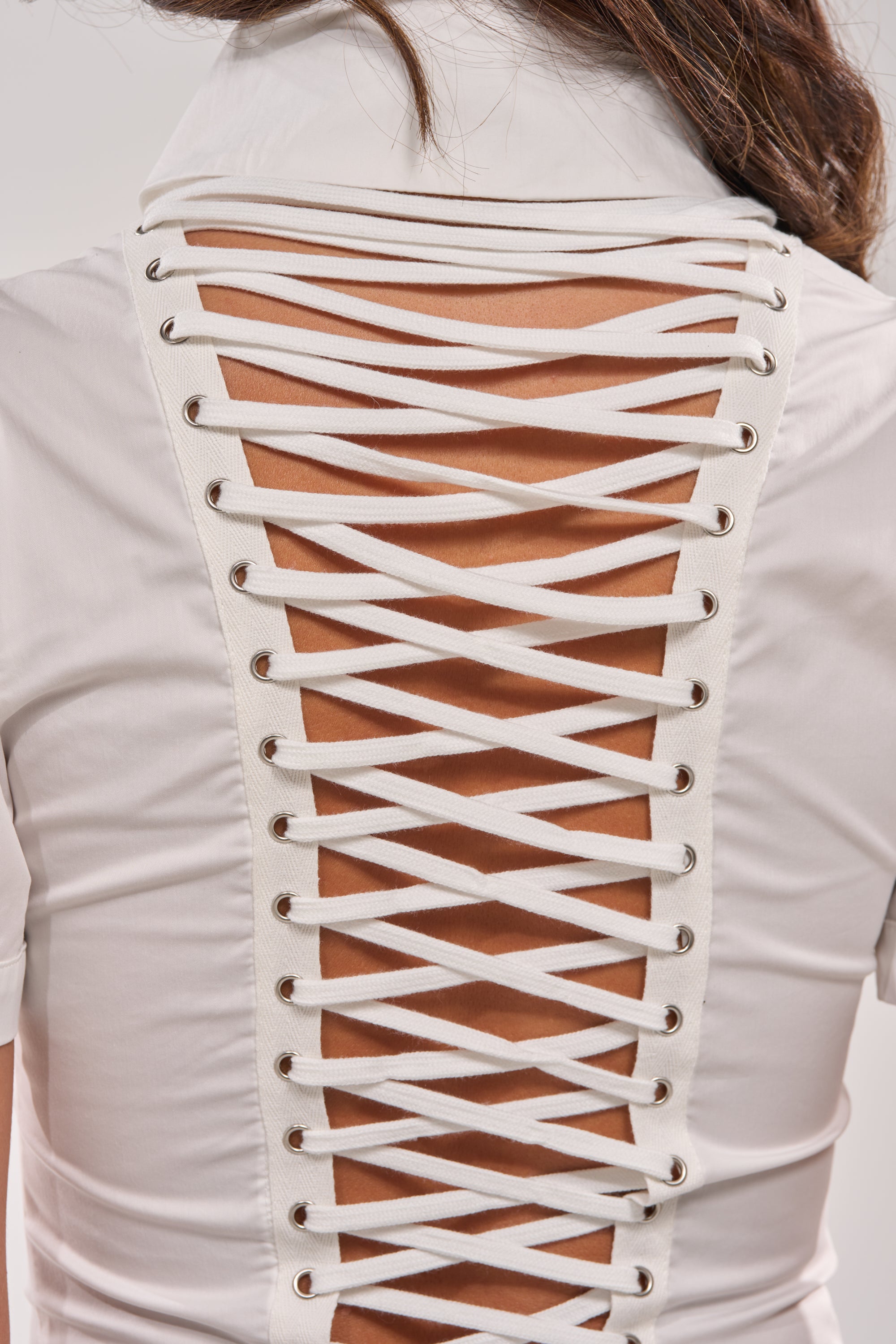 A person wears the PRETTY PLEASE BUTTON DOWN BLOUSE, a white lace-up top with an open back and crisscross laces threaded through metal eyelets, revealing skin underneath. 8