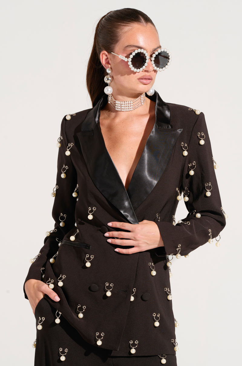 A person wearing the DO AS I PLEASE TRINKET BLAZER, embellished with faux pearl charms and featuring a shiny black lapel. They complement the look with oversized sunglasses studded with pearls, pearl earrings, and a choker necklace. Their hair is styled in a sleek ponytail.