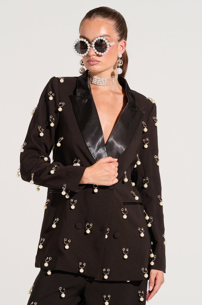 A woman in a fashionable DO AS I PLEASE trinket blazer with pearl accents strikes a confident pose. She accessorizes with oversized, round sunglasses embellished with pearls and a faux pearl choker, while her hair is tied back. The blazer boasts a satin lapel, enhancing the elegance of her outfit.