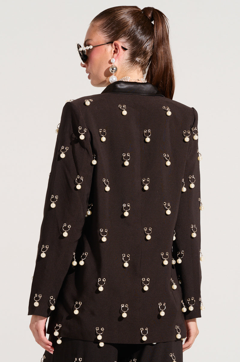 A person with long hair styled in a ponytail, adorned with sunglasses and earrings, is facing away. They are wearing the "Do As I Please Trinket Blazer" featuring faux pearl charms and matching pants. The background is plain and light-colored.