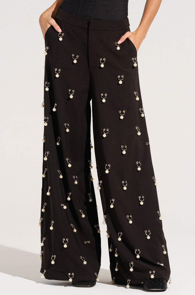A person models the DO AS I PLEASE TRINKET TROUSER, showcasing its high-rise wide-leg design and metallic embellishments. The trousers are adorned with numerous evenly spaced, shiny circular designs that shimmer like faux pearl charms as the individual stands casually with hands in pockets.