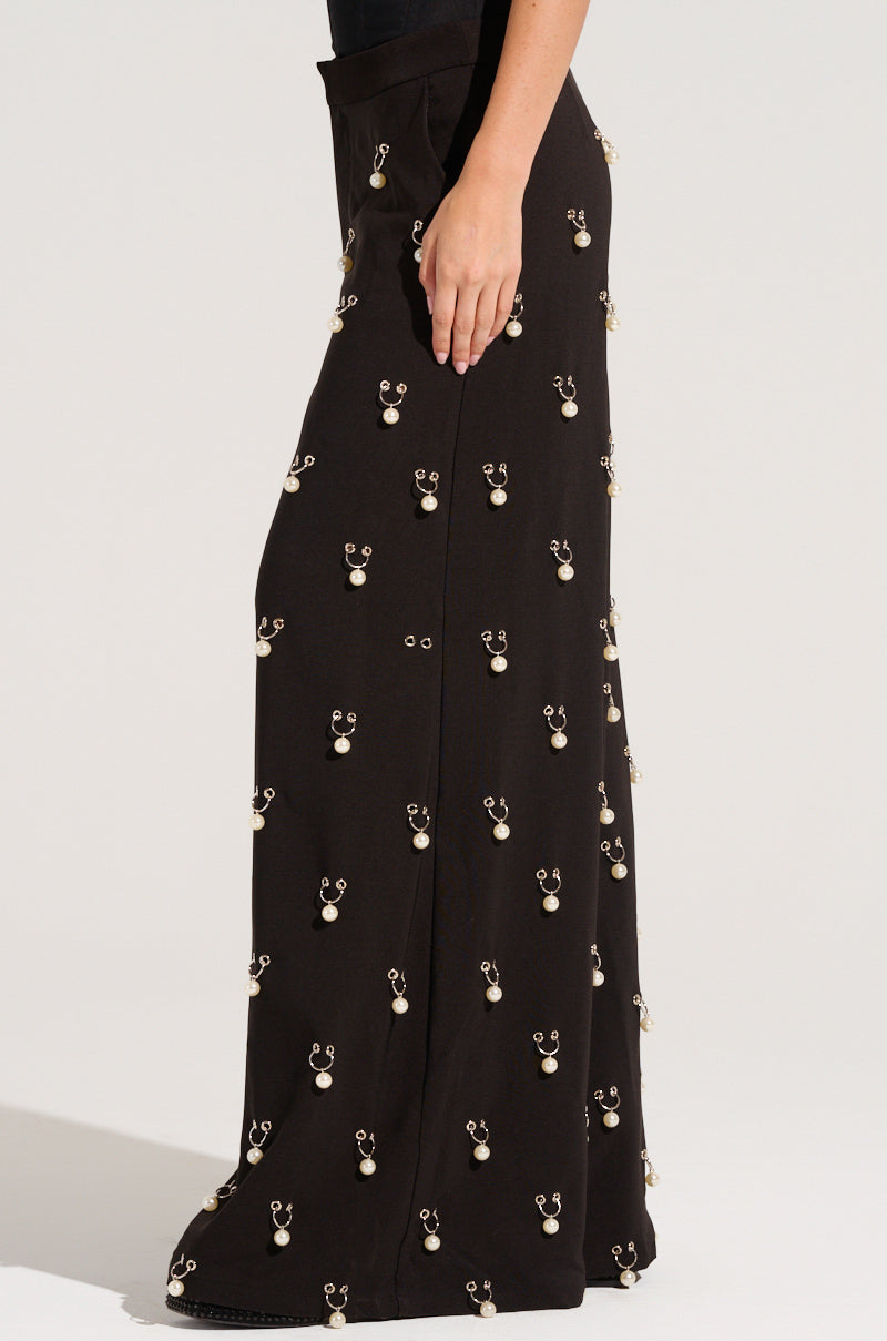 A woman stands in profile wearing the DO AS I PLEASE TRINKET TROUSER, which are high-rise, wide-leg trousers featuring a pattern of small, light-colored floral embellishments and subtle faux pearl charms.