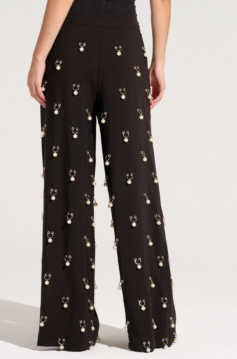 The woman is seen from behind, showcasing the DO AS I PLEASE TRINKET TROUSER in chic black. These wide-leg trousers are embellished with dangling silver hoops and faux pearl charms, offering a high-rise fit adorned with metallic decorations throughout.