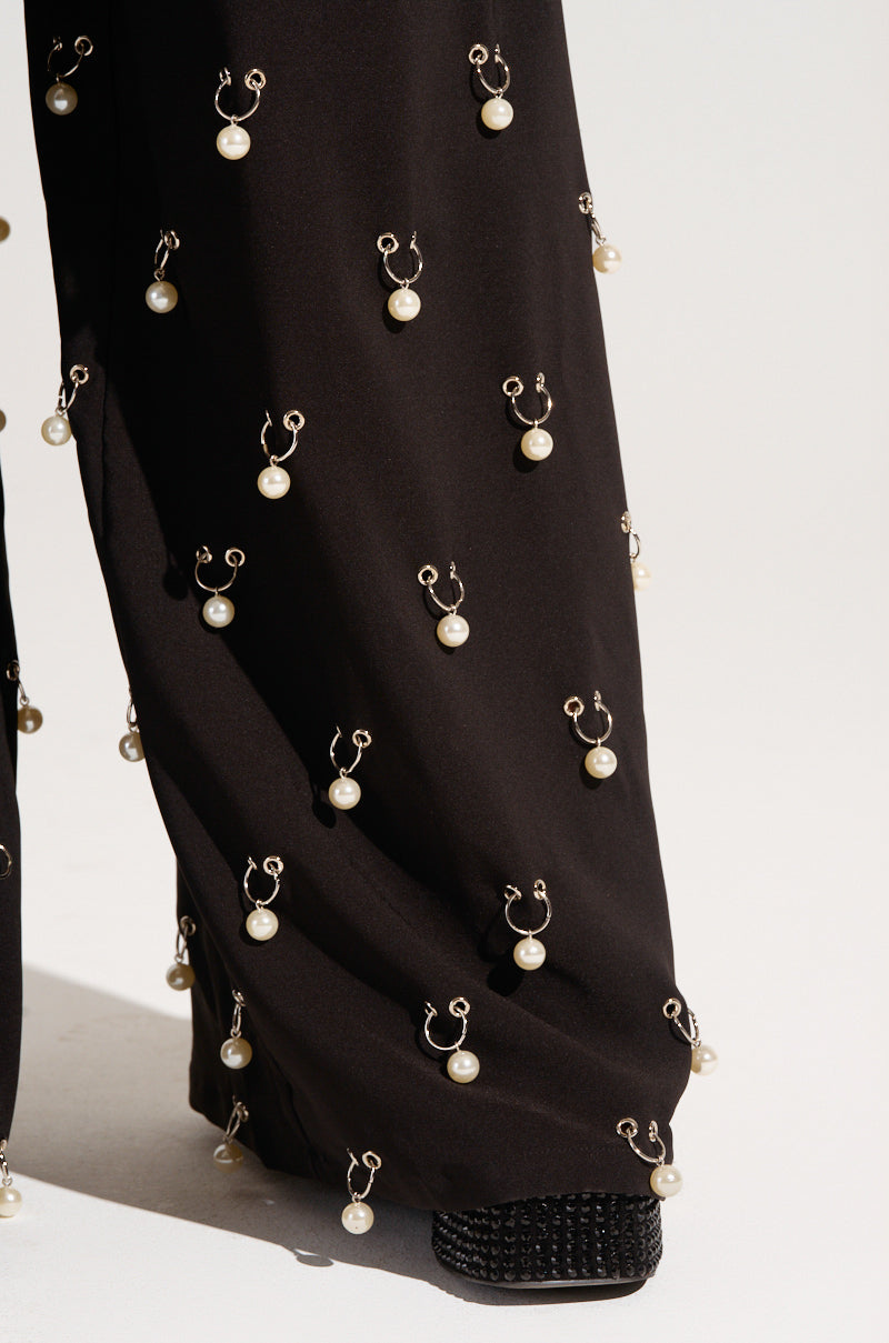 The DO AS I PLEASE TRINKET TROUSER features black, wide-leg design accentuated with faux pearl charms and small hooks that form a distinctive decorative pattern. These high-rise trousers are styled with woven black shoes peeking out from beneath the hem, set against a plain, neutral background.