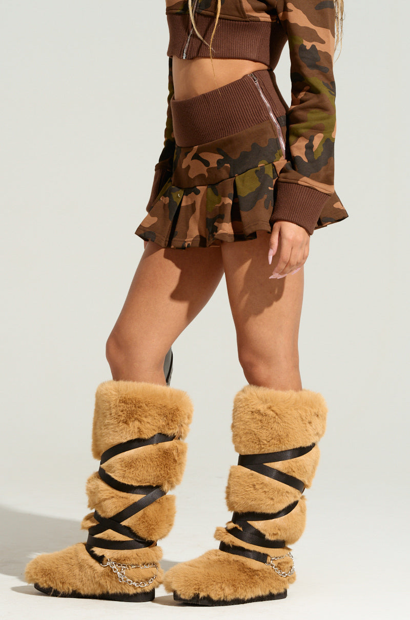 The individual confidently flaunts an AKIRA Label camo print crop top paired with the LOVE ME LIKE THAT MINI SKIRT. Complemented by fur knee-high boots adorned with black straps and chains, this bold fashion statement stands out against a plain light background.