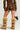 A person wearing the LOVE ME LIKE THAT MINI SKIRT in camo print paired with a brown top. They have on tan, furry boots featuring black crisscross straps and chain details, and they're carrying a small black handbag with a chain strap, exuding an AKIRA Label style.