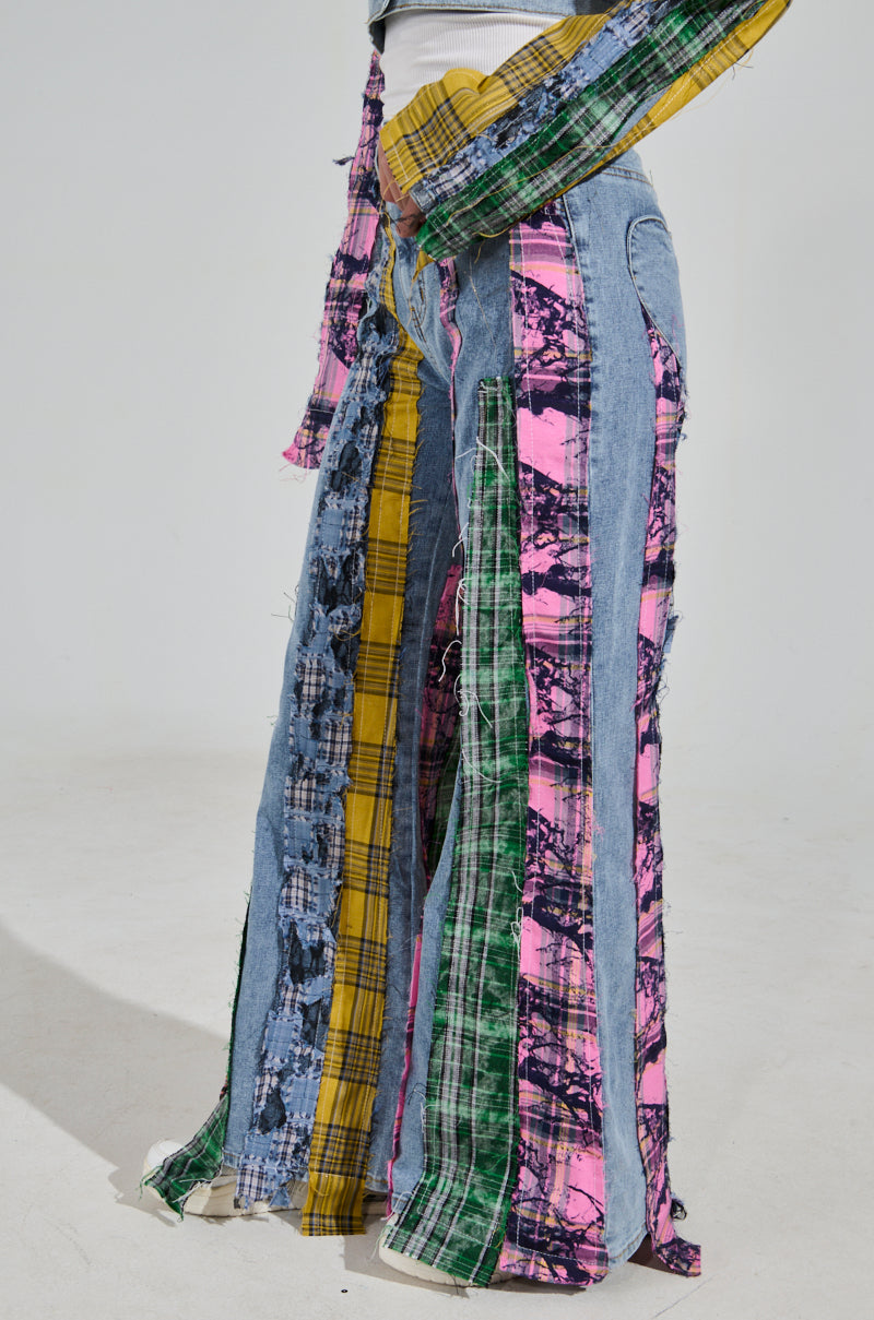 A side view of someone wearing the MAKING WAVES PATCHWORK FLARE DENIM shows high-rise pants with vertical strips of colorful plaid and tie-dye denim, offering a bold, eclectic style reminiscent of distressed plaid flannel, against a plain background.