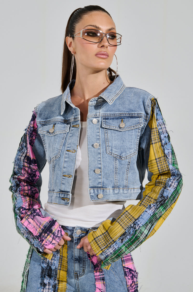 A person wears the MAKING WAVES PATCHWORK DENIM JACKET with colorful sleeves, standing confidently. With long hair, sunglasses, hoop earrings, and denim pants, their eye-catching look is highlighted against a plain background.