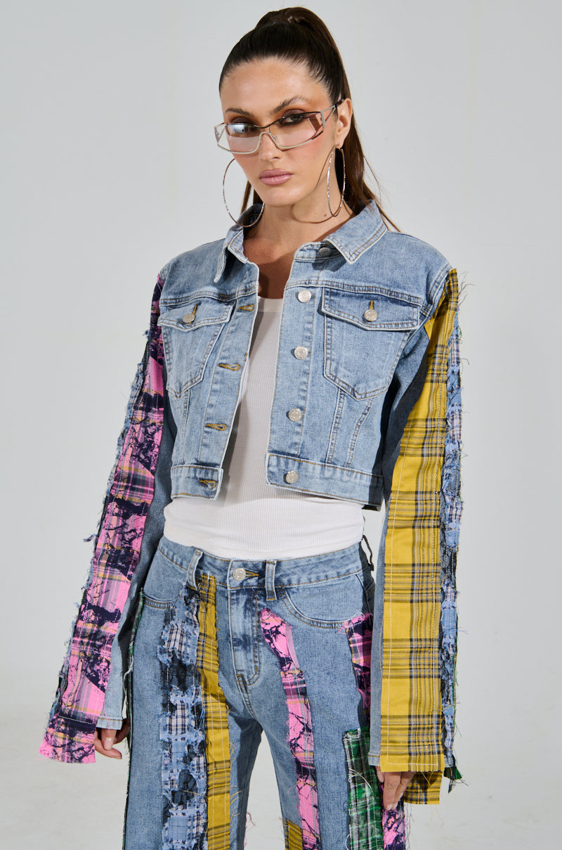 A woman showcases a chic style wearing the MAKING WAVES PATCHWORK DENIM JACKET with matching jeans featuring colorful plaid patches in pink, yellow, and green. Her long hair is tied back, accented by large sunglasses and a white top under the jacket.