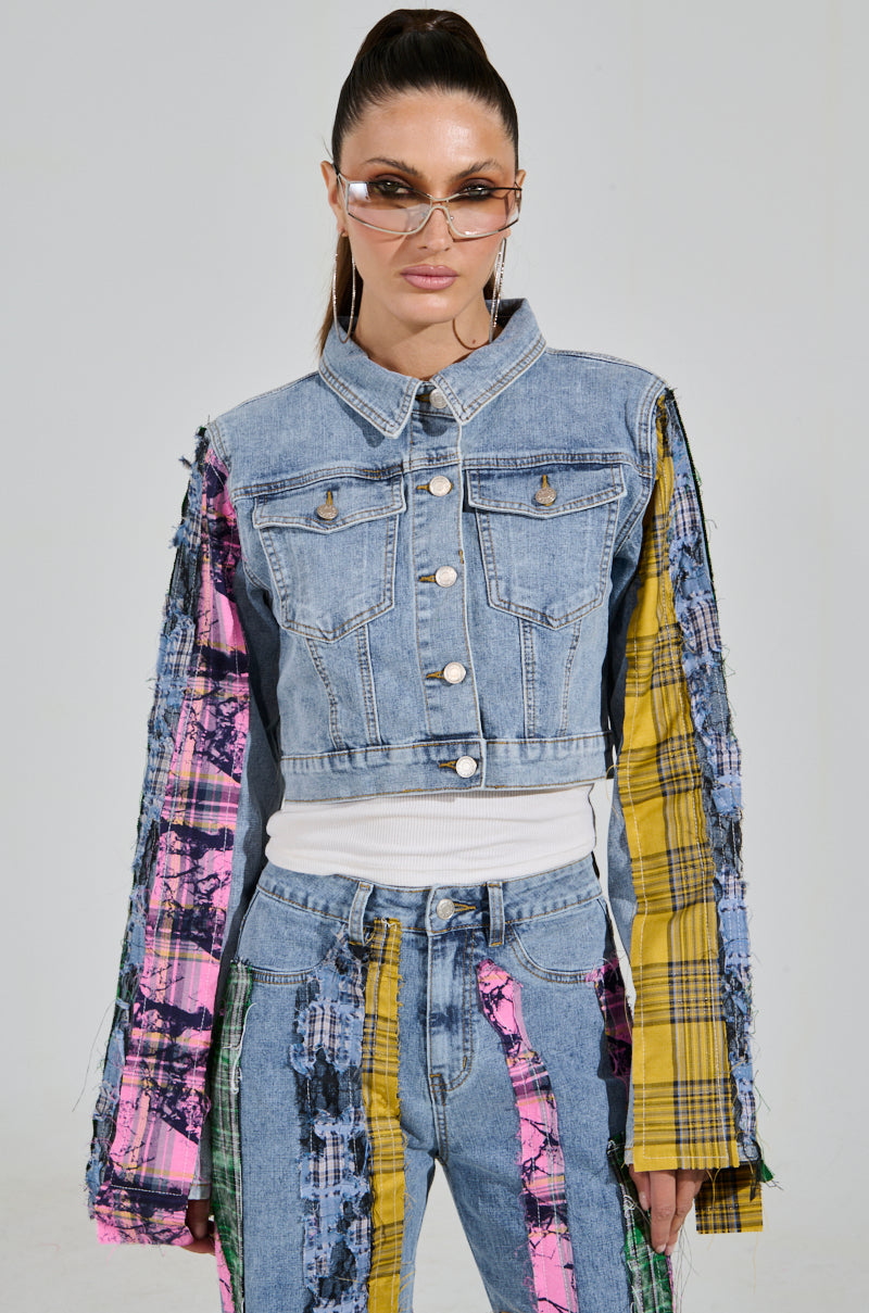 Wearing sunglasses and the MAKING WAVES PATCHWORK DENIM JACKET, a person flaunts a cropped boxy silhouette with colorful plaid and abstract patterns. The long-sleeve jacket pairs perfectly with matching pants against a plain gray background.