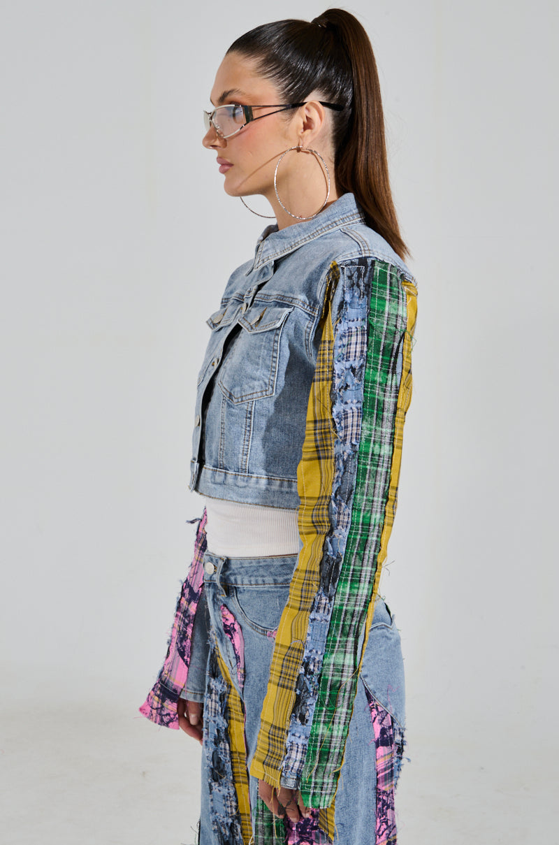 A person with long hair in a ponytail is wearing glasses, hoop earrings, and a unique outfit featuring the MAKING WAVES PATCHWORK DENIM JACKET with colorful plaid sleeves, complemented by similar jeans. The cropped boxy silhouette stands out against a plain white background.