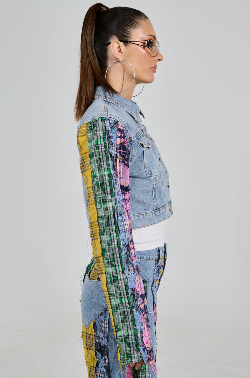 A person with long hair in a ponytail, wearing sunglasses and large hoop earrings, is dressed in the MAKING WAVES PATCHWORK DENIM JACKET with colorful plaid patches, paired with matching patterned pants. They stand sideways against a plain background.