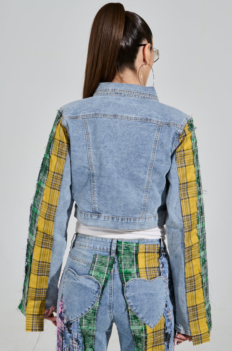 A person with a high ponytail wears the MAKING WAVES PATCHWORK DENIM JACKET, featuring a cropped boxy silhouette and yellow and green plaid sleeves, along with jeans adorned with heart-shaped patches. They face away from the camera against a neutral background.