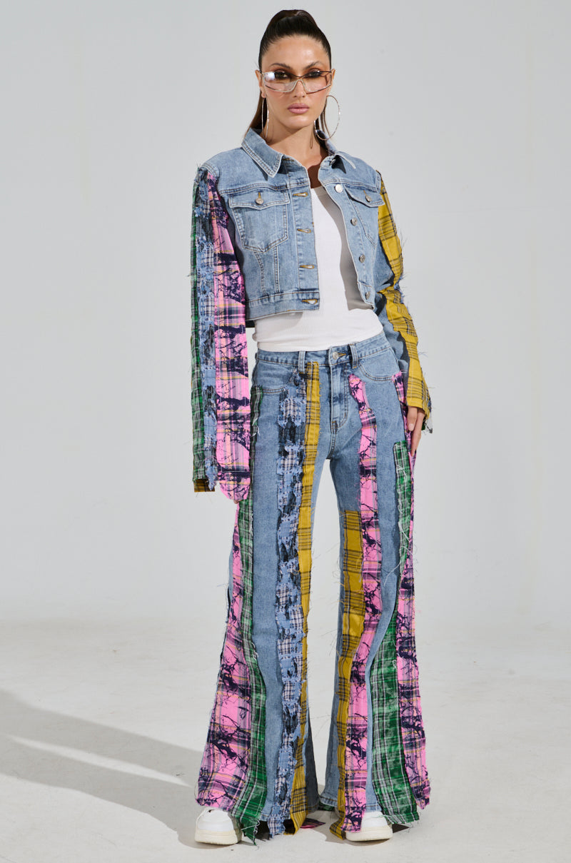A person wearing the MAKING WAVES PATCHWORK DENIM JACKET with colorful striped patterns complements their flared pants. They sport a cropped boxy silhouette, finishing the look with sunglasses, hoop earrings, and white sneakers, standing against a plain background.