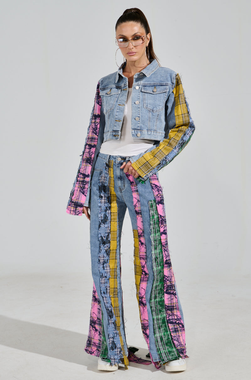 Against a plain backdrop, an individual sports the MAKING WAVES PATCHWORK DENIM JACKET with pink and yellow plaid sleeves. The matching pants feature multicolored panels and bleach patterns. Glasses enhance the outfit's distinctive style with distressed plaid flannel details.