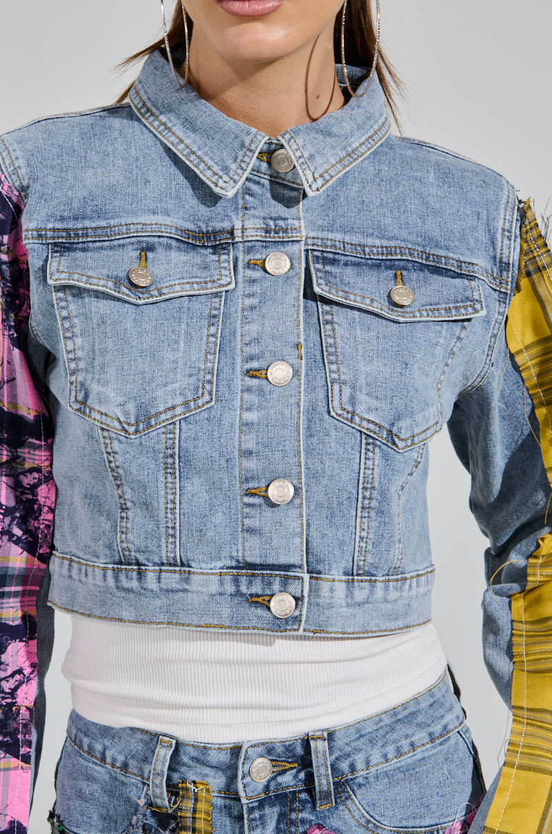 The woman wears the MAKING WAVES PATCHWORK DENIM JACKET with a cropped boxy silhouette, featuring plaid and abstract designs in pink and yellow, paired with matching denim pants. The outfit includes metallic buttons and a white top underneath.