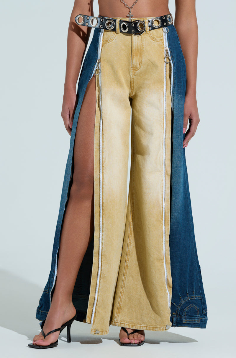 Someone wearing SHE CAN DO BOTH MIXED MEDIA DENIM JEANS, featuring a light tan front panel, zippers, exposed sides, two-tone wide-leg silhouette, styled with a black studded belt and black heeled sandals.