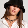 A woman with straight brown hair wears the RUCHED BABE SATIN BUCKET HAT and a white blouse. She accessorizes with a black choker necklace featuring small dangling cross charms, adding a touch of festival accessory flair. The background is plain and light-colored.