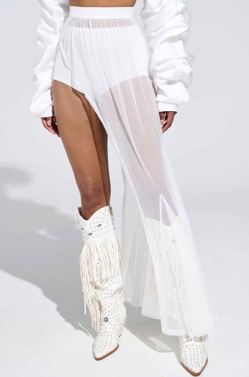 A person poses against a plain backdrop, wearing the ULTIMATE LAYER SKIRT—a sheer white asymmetrical piece over high-waisted shorts. The look is completed with voluminous sleeves and white cowboy boots featuring fringe and studs, all enhanced by an elastic waistband for comfort.