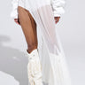 A person poses against a plain backdrop, wearing the ULTIMATE LAYER SKIRT—a sheer white asymmetrical piece over high-waisted shorts. The look is completed with voluminous sleeves and white cowboy boots featuring fringe and studs, all enhanced by an elastic waistband for comfort.
