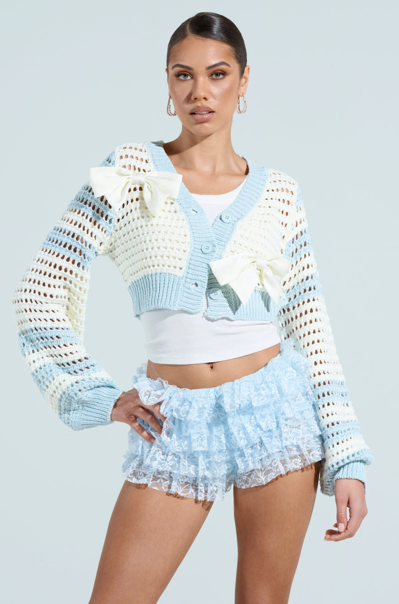 A woman confidently poses in the FLOWER GIRL CARDIGAN, a light blue and white cropped cardigan with oversized satin bow accents, over a white top. She's wearing light blue ruffled shorts and hoop earrings, with her hair tied back against a plain light background.
