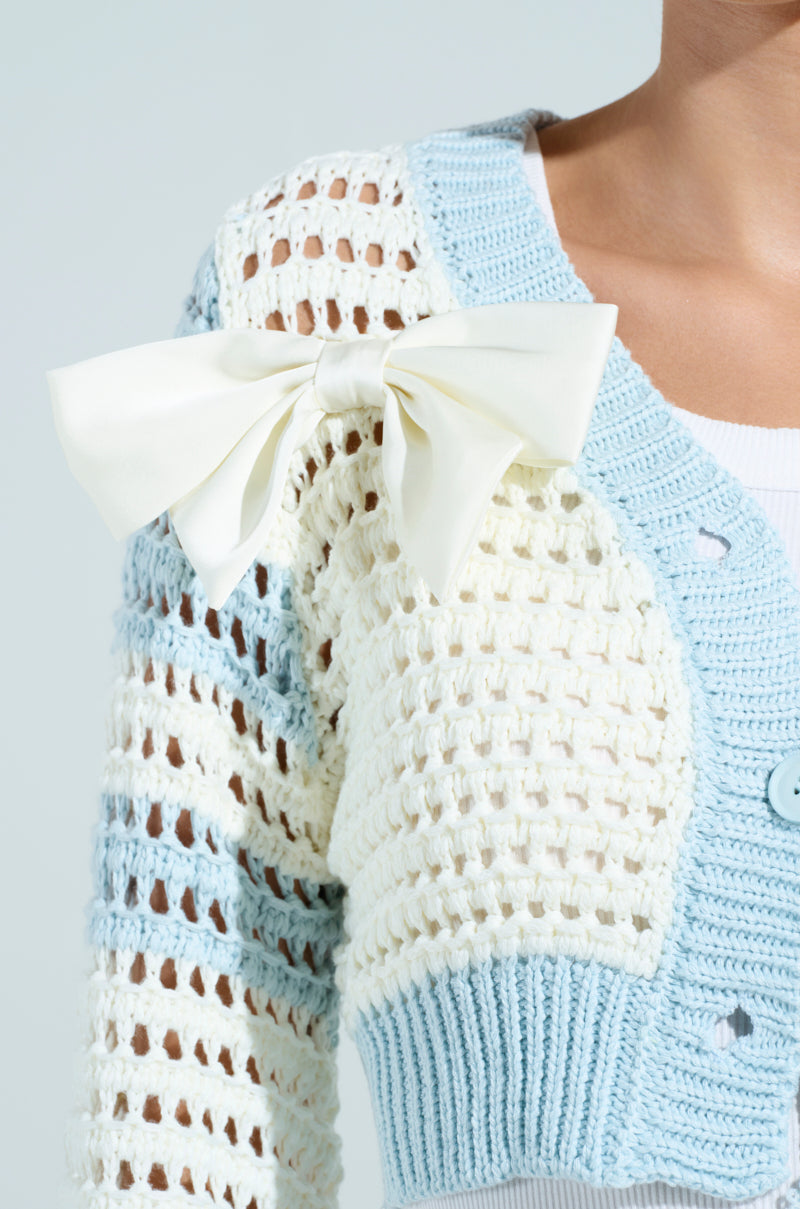 A person wears a FLOWER GIRL CARDIGAN with alternating blue and white stripes, featuring a large white bow on the shoulder and blue buttons, stylishly layered over a white top.