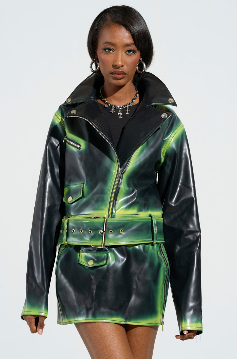 A person wearing the LIGHTS OUT PAINTED SEAM MOTO jacket and skirt in black and neon green faux leather, with a black shirt underneath. They have straight dark hair and wear a necklace with cross pendants. The background is a simple, light color.