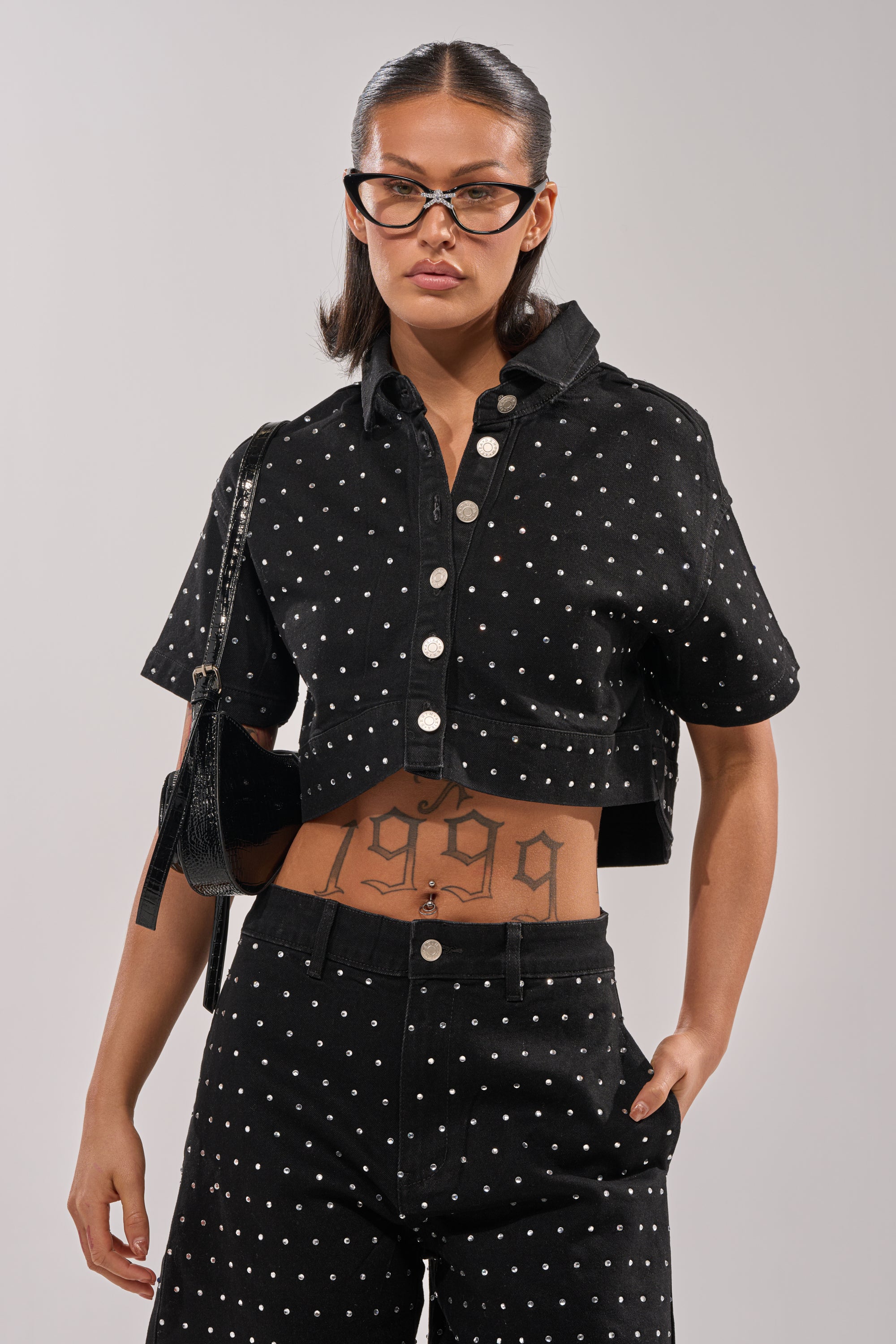 Wearing the ITS GIVING DENIM RHINESTONE BUTTON DOWN and matching polka dot pants, black cat-eye glasses, and a shoulder bag, this person shows off a "1999" stomach tattoo. 0