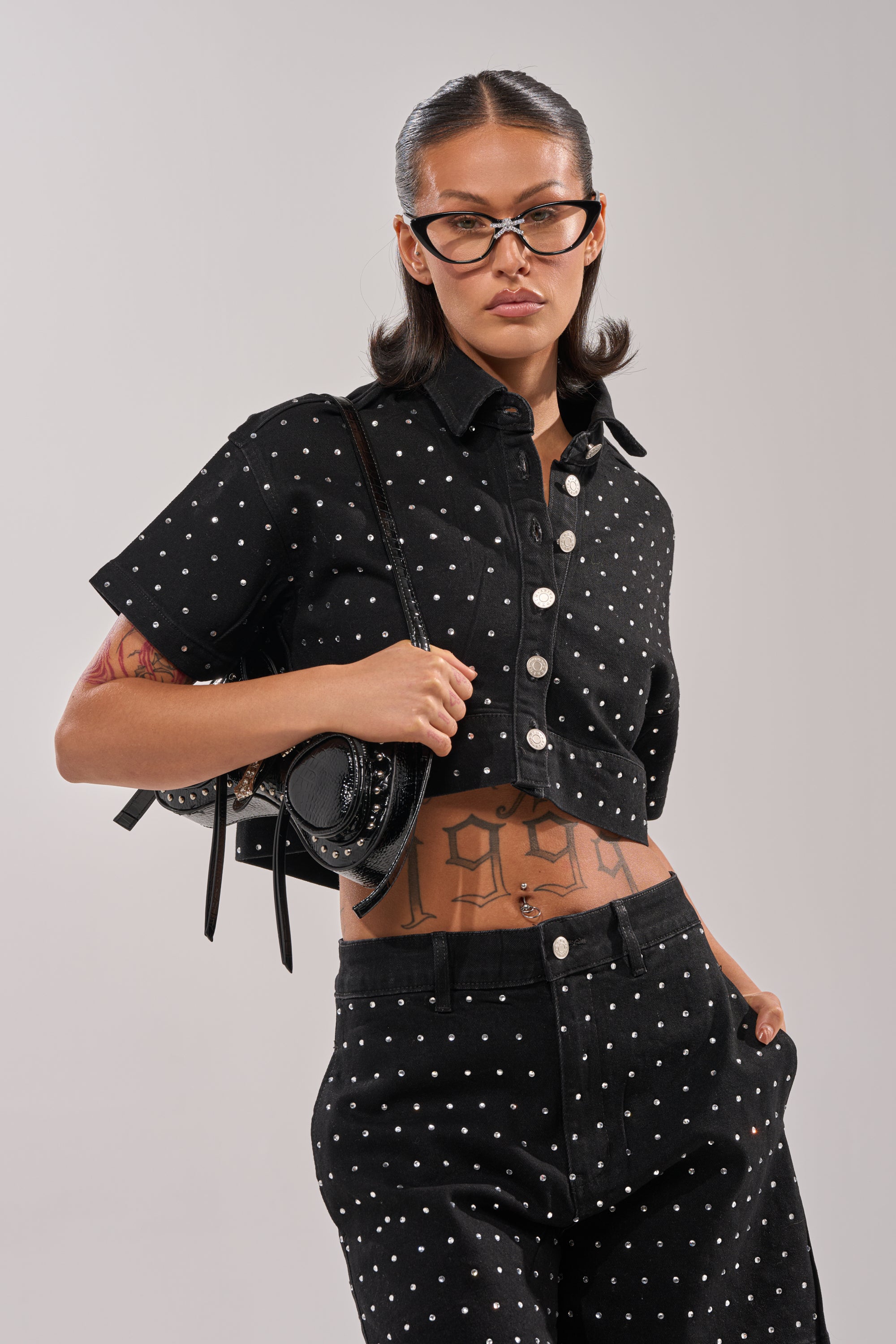 Wearing the ITS GIVING DENIM RHINESTONE BUTTON DOWN, a person stands confidently with one hand in their pocket, matching pants, large glasses, a small black backpack, and a “1998” tattoo showing beneath their cropped denim shirt. 1