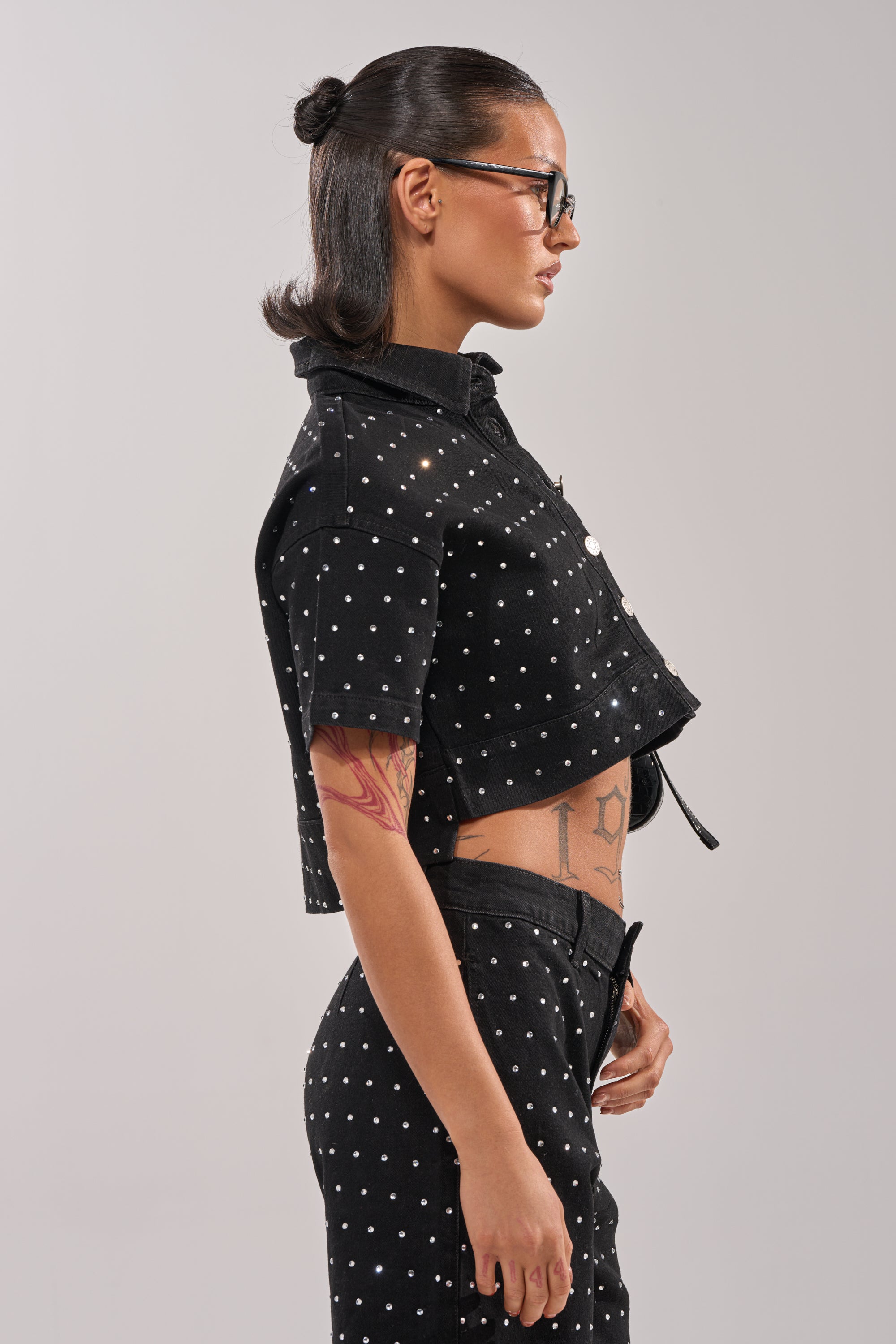 A person with dark hair in a bun and glasses poses in profile, wearing the ITS GIVING DENIM RHINESTONE BUTTON DOWN shirt with black polka dots and matching pants. A red tattoo is visible on their arm against a plain light gray background. 2
