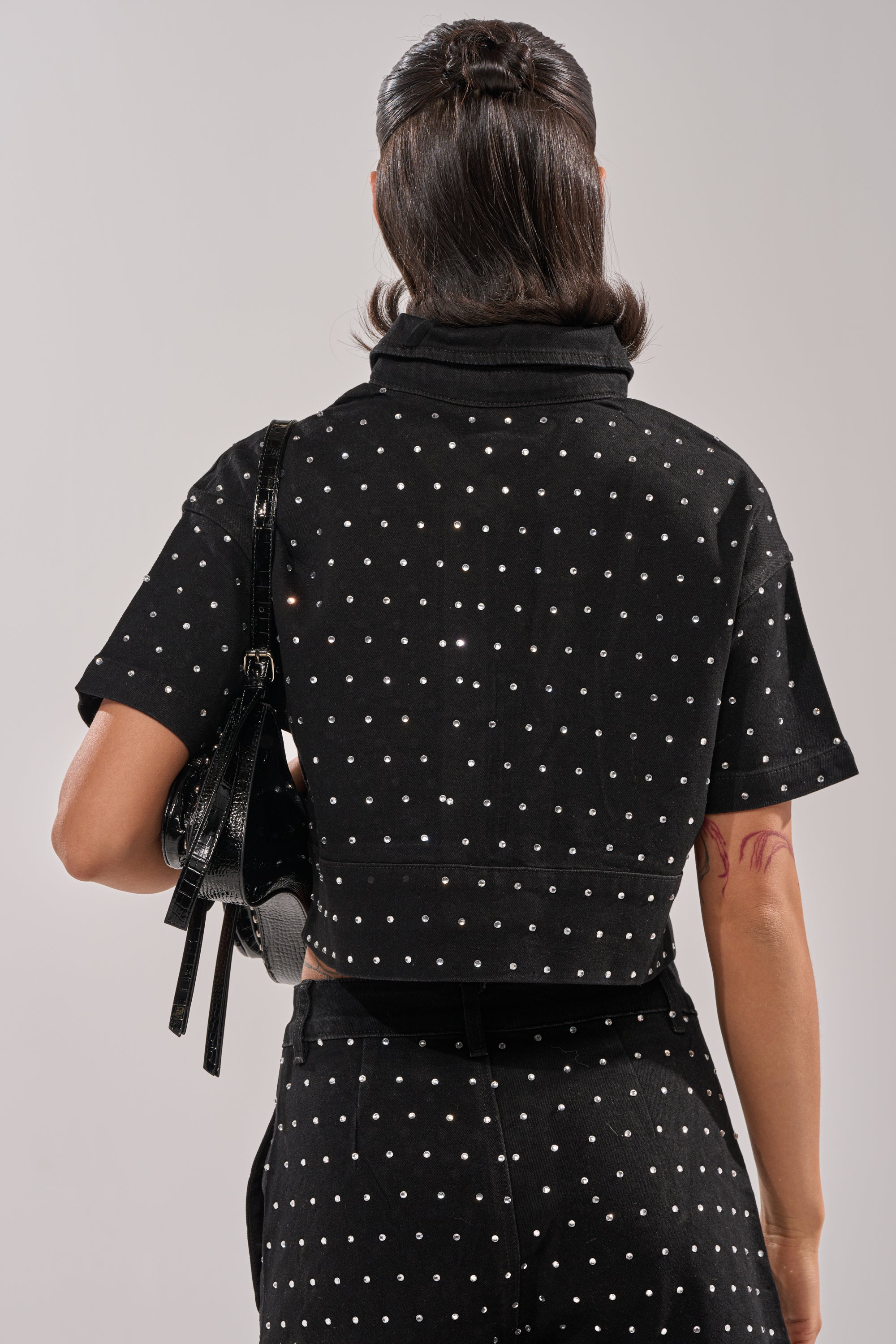 A person faces away, wearing the ITS GIVING DENIM RHINESTONE BUTTON DOWN—a cropped, polka-dot denim shirt with matching pants—and carries a shiny black handbag on their shoulder against a plain, neutral background. 3