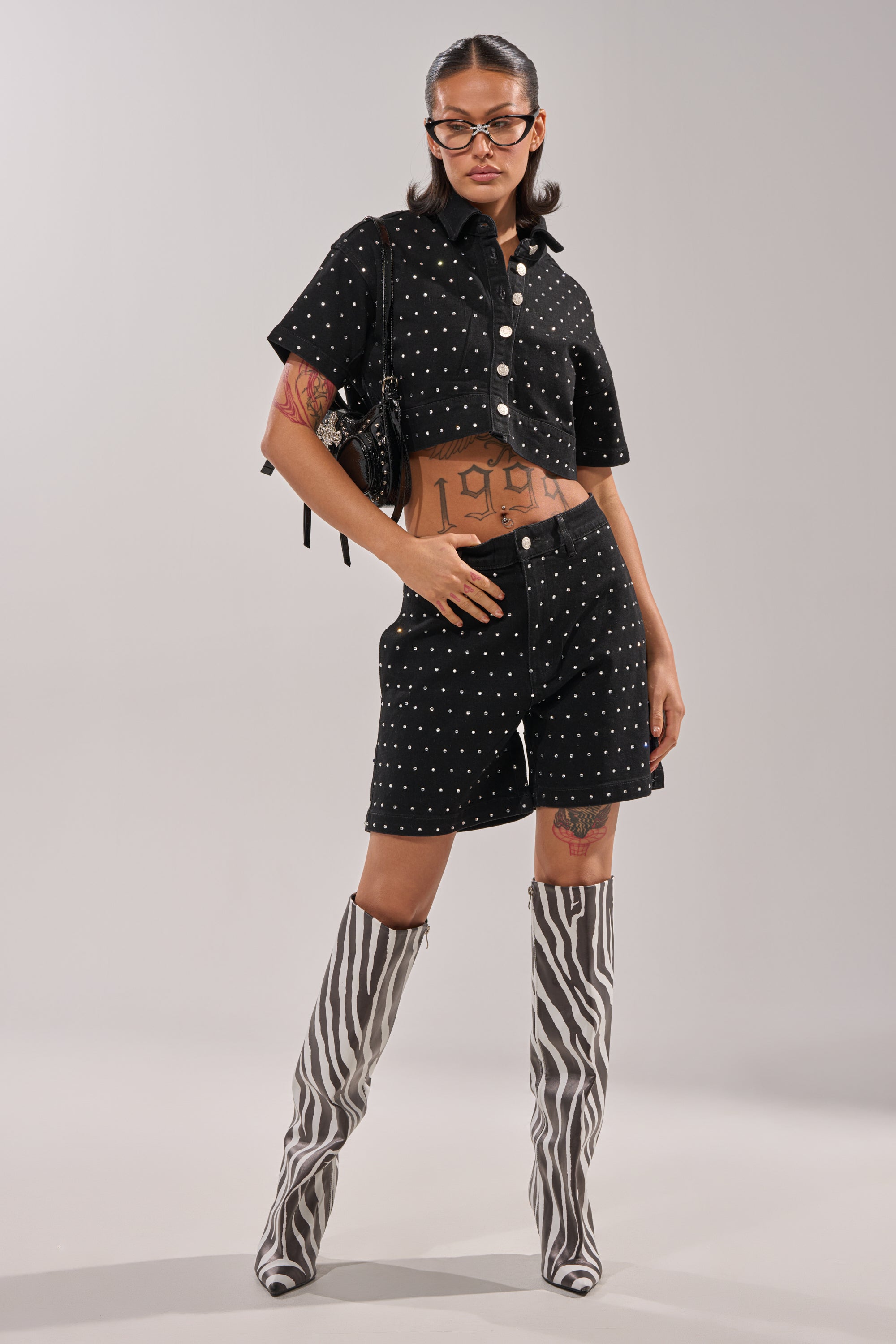 A woman stands confidently in the ITS GIVING DENIM RHINESTONE BUTTON DOWN top and matching shorts, paired with zebra-print knee-high boots, glasses, a black backpack, and showcasing tattoos on her stomach and thigh. 5