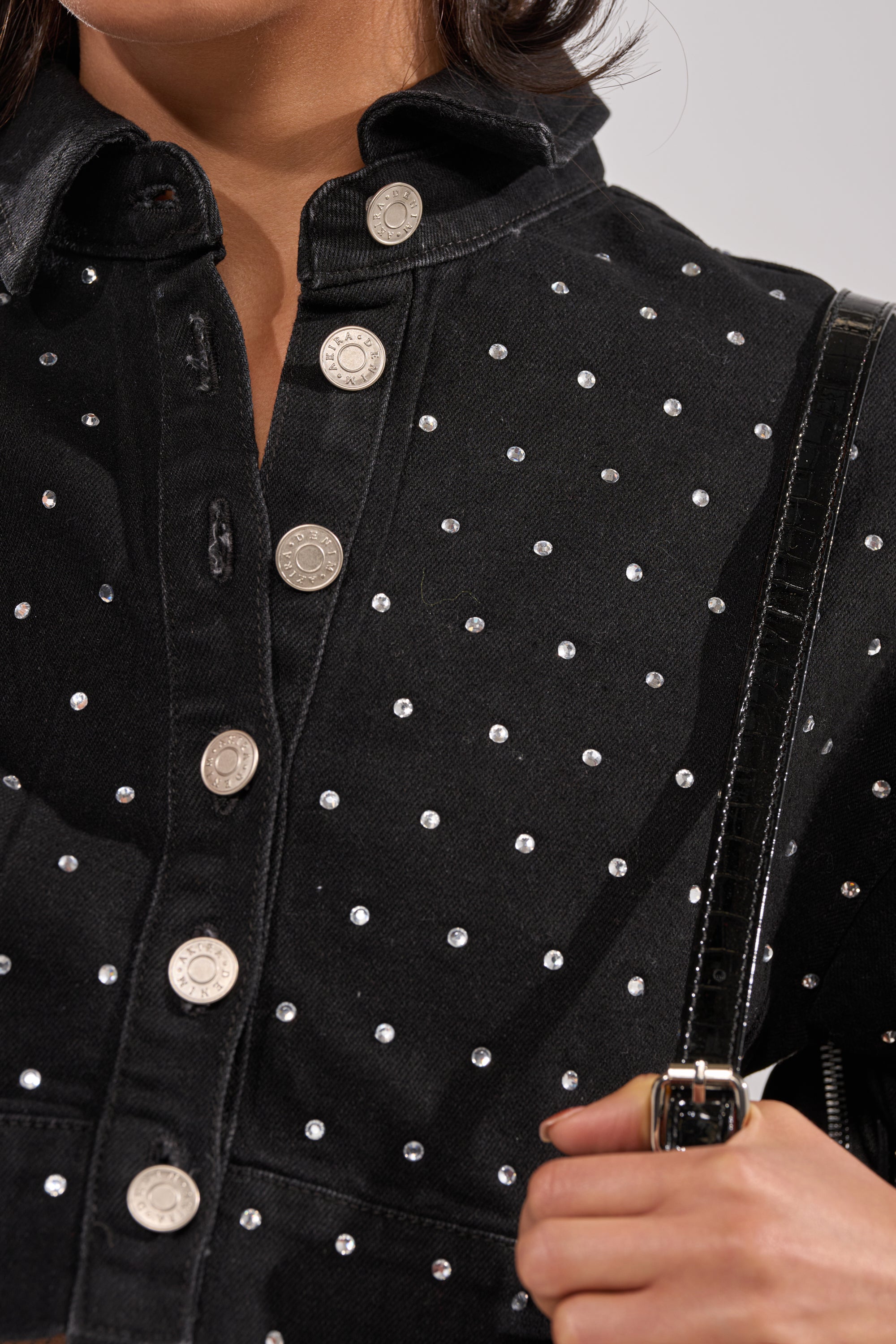 Close-up of a person wearing the ITS GIVING DENIM RHINESTONE BUTTON DOWN, a black-wash denim top with silver studs and buttons, carrying a black strap purse with a gold buckle on their shoulder. 6