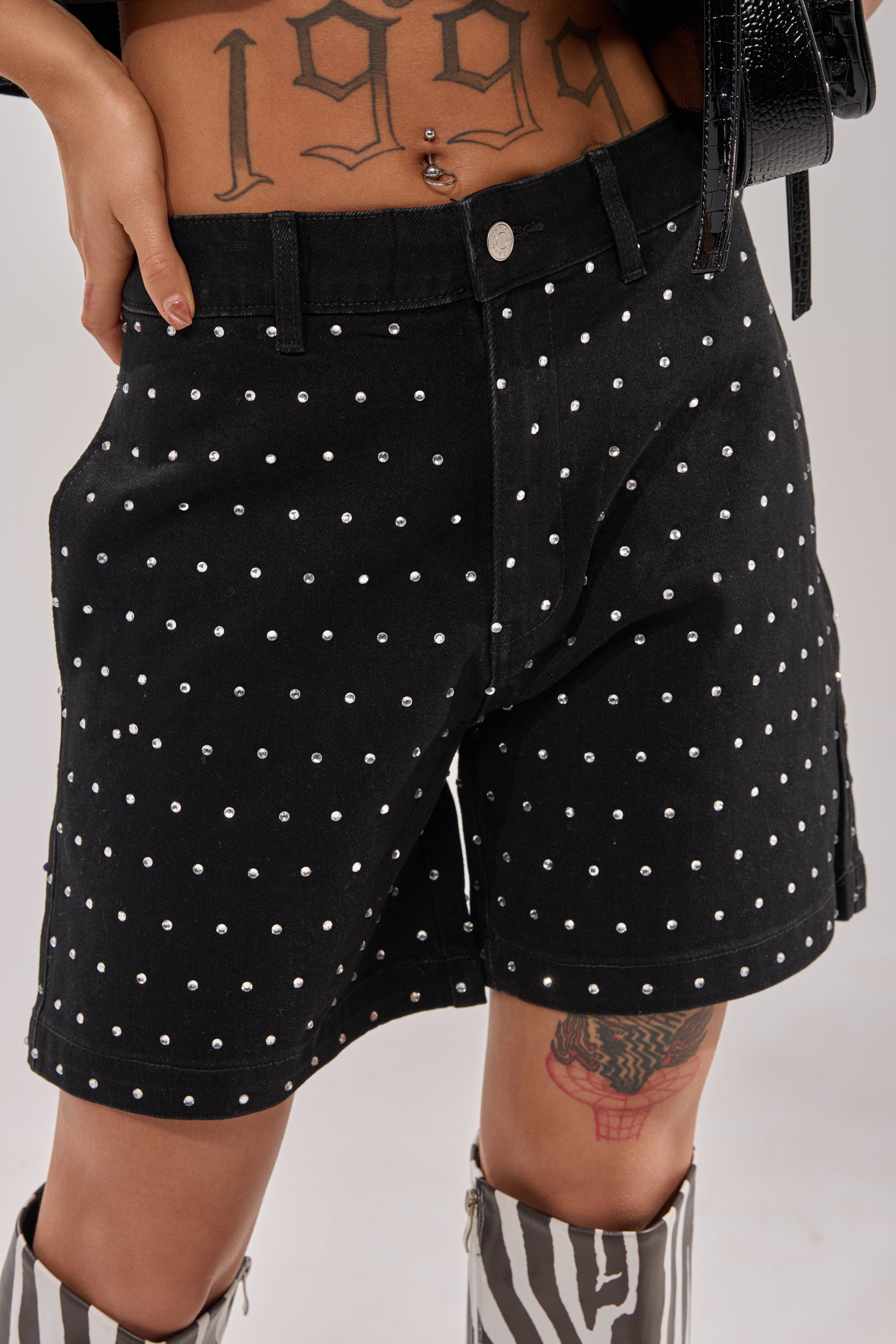Wearing the ITS GIVING BERMUDA SHORT in black polka-dot, this person shows off a "1999" lower abdomen tattoo, a thigh tattoo, zebra-print boots, and carries a black handbag while posing with a hand on their hip. 0