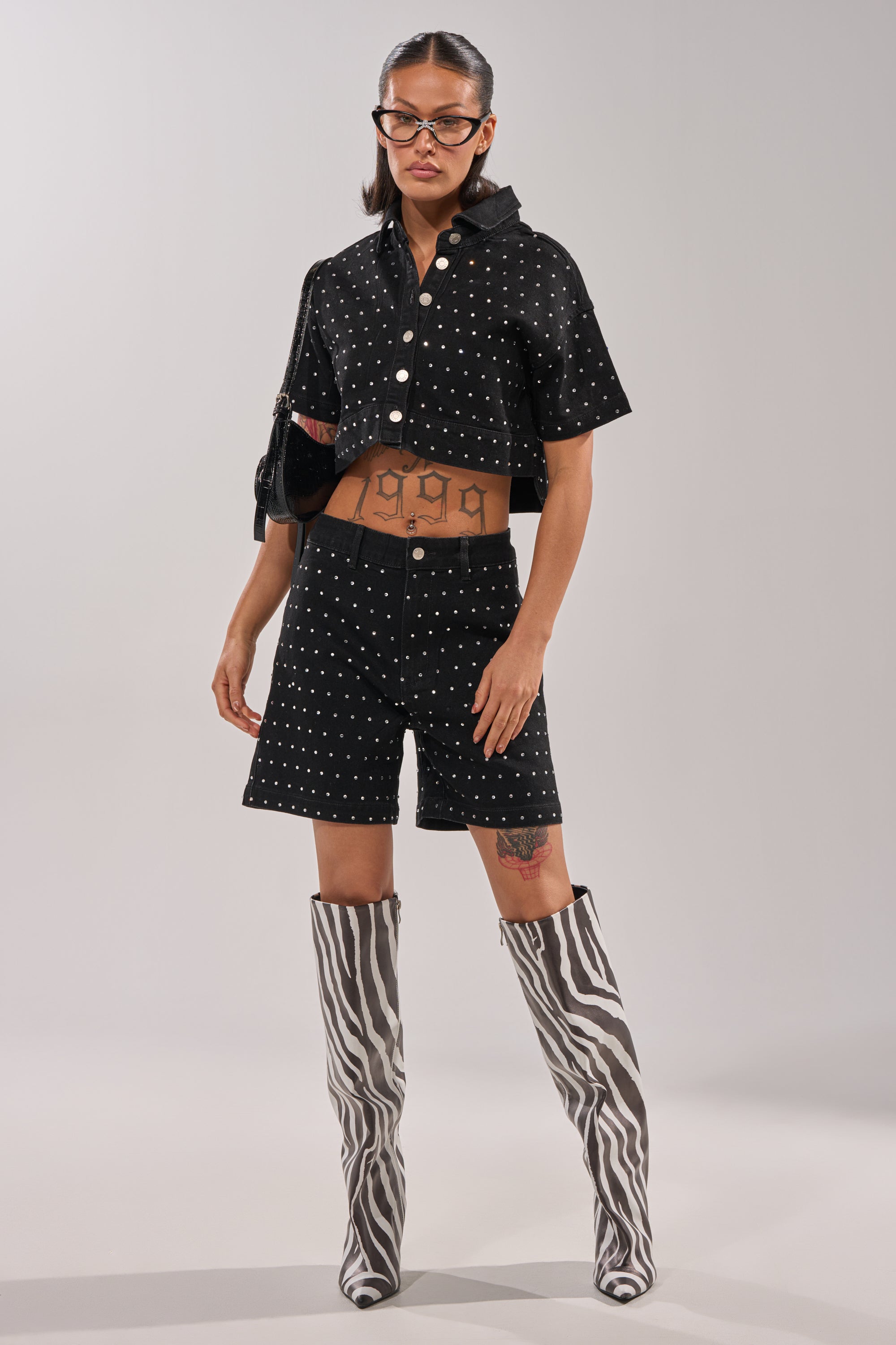 A woman in the ITS GIVING BERMUDA SHORT, a black studded short-sleeve jacket, zebra-print knee-high boots, black purse, glasses, and hoop earrings stands against a light background, showing tattoos on her legs and stomach. 5