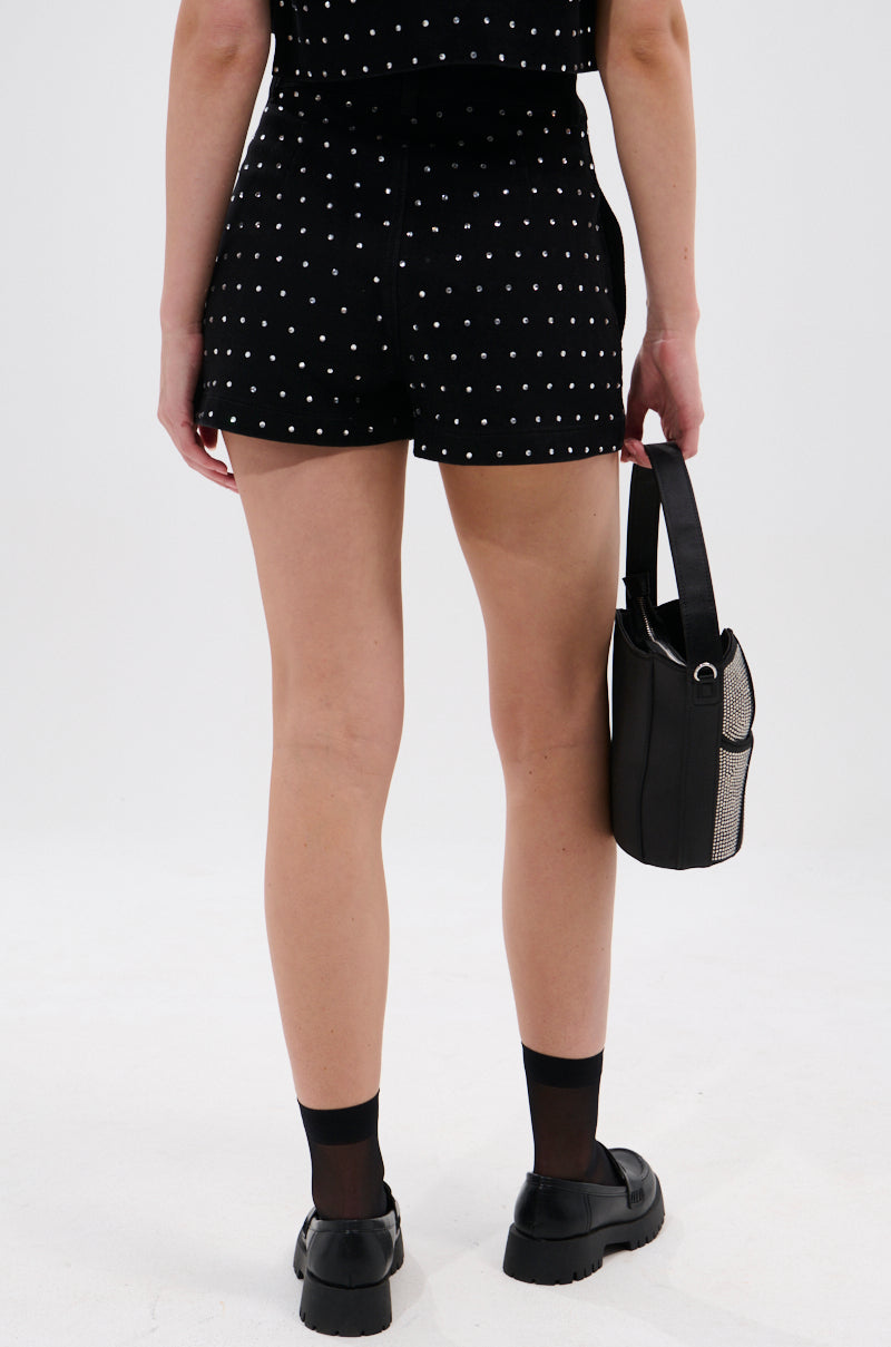 A person stands facing away, wearing the ITS GIVING DENIM RHINESTONE SHORT with black socks, black shoes, and a small black handbag against a plain white background—perfect summer wardrobe inspiration. 3