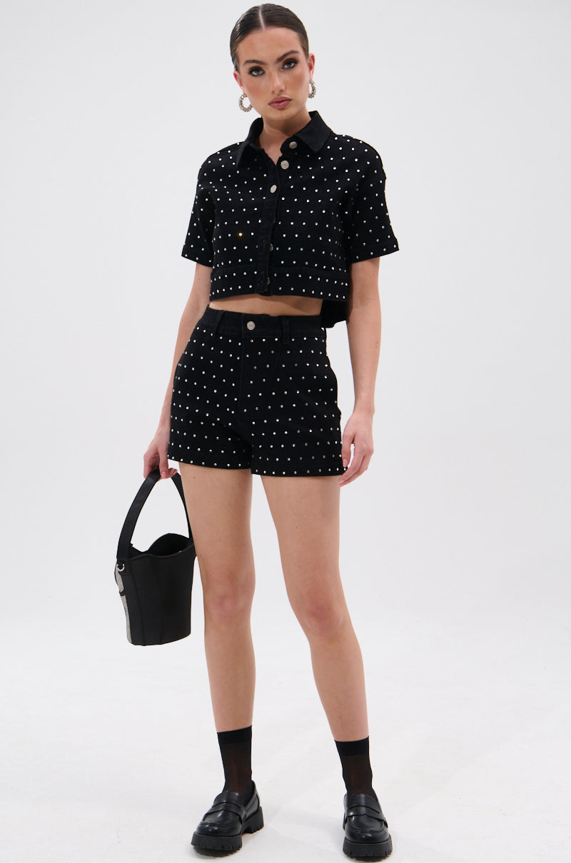 A woman stands against a white background wearing the ITS GIVING DENIM RHINESTONE SHORT—a black, high-waisted short with sparkling rhinestone details—paired with a matching crop top, black handbag, and loafers for an effortless summer look. 5