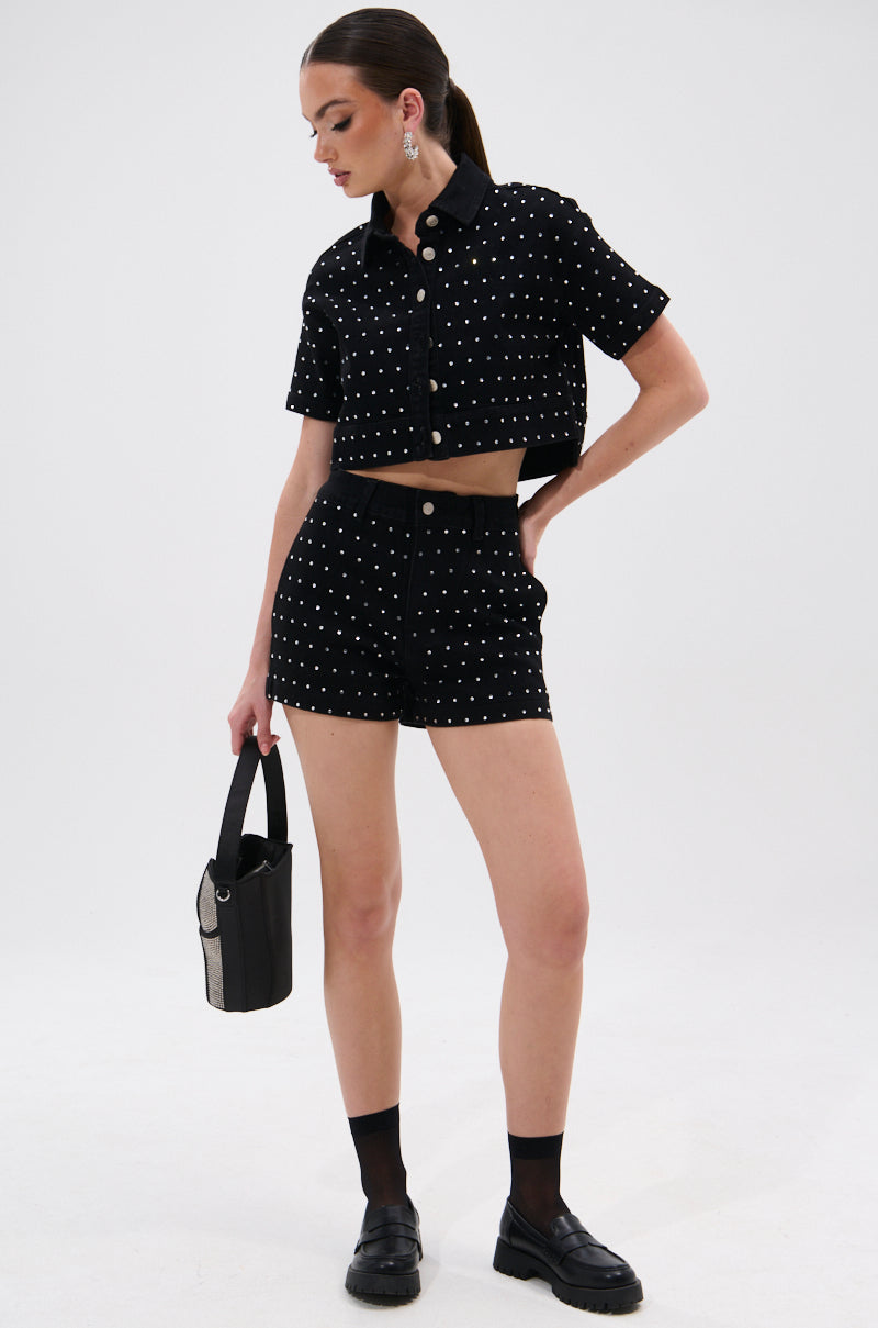 A woman models the ITS GIVING DENIM RHINESTONE SHORT—a chic, high-waisted short with dazzling rhinestones. She completes the look with a black handbag, loafers, socks, and statement earrings for effortless summer style. 6