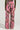 A person is seen from behind wearing the STAND OUT TO FIT IN CARGO PANTS, featuring a pink camouflage pattern with abstract tree branch designs and a wide leg silhouette. They have paired the pants with a green long-sleeve top, standing upright against a plain background.