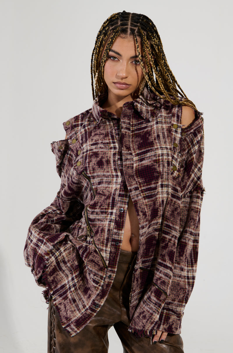 A person with braided hair poses confidently in an oversized DRIFTER FLANNEL featuring purple plaid, cut-out shoulders, and distressed detailing, paired with brown pants. They have one hand in their pocket against a plain background.