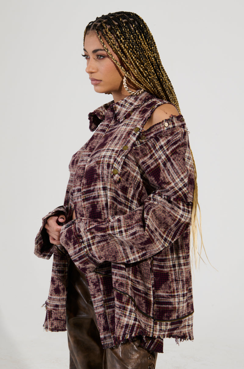 A person with long braided hair is shown in profile against a plain background, wearing the DRIFTER FLANNEL, a distressed plaid shirt featuring cut-out shoulders and an oversized fit. They complement the grunge-style shirt with dark pants and large hoop earrings.