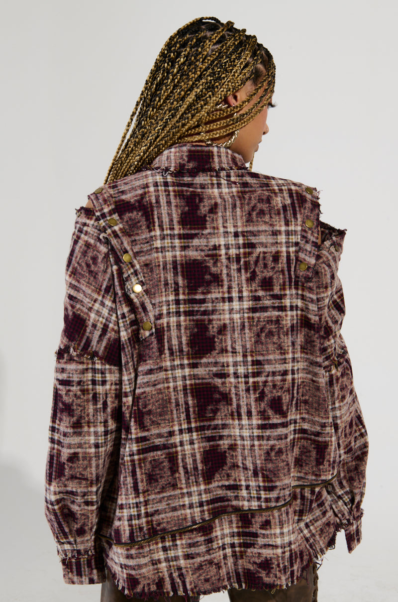 A person with braided hair is facing away from the camera, highlighting the DRIFTER FLANNEL in purple and beige plaid with distressed detailing. This grunge flannel boasts an oversized fit, blending seamlessly into the neutral background.