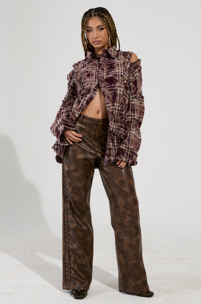 A person with braided hair poses confidently, showcasing an oversized fit in the DRIFTER FLANNEL top paired with brown leather pants featuring lace detailing. The plain white background accentuates the outfit's unique textures and design.