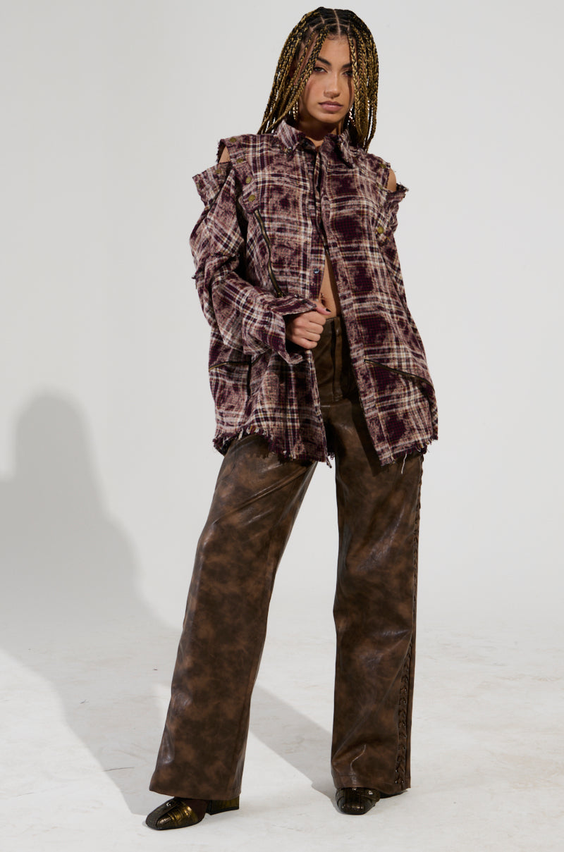 A person with braided hair stands confidently in the DRIFTER FLANNEL featuring distressed plaid detailing and pairs it with loose, brown leather pants. The plain background highlights their bold fashion style.